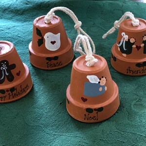 May include: Four terracotta pot bells with painted holiday themes. The bells are decorated with a gingerbread man, a dove, two angels, and a single angel. The bells are hung with white twine and have the words "Happy Holidays", "Peace", "Friends", and "Noel" written on them.