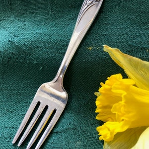 May include: A silver fork with a floral design on the handle. The fork is lying on a green tablecloth with a yellow daffodil flower to the right.