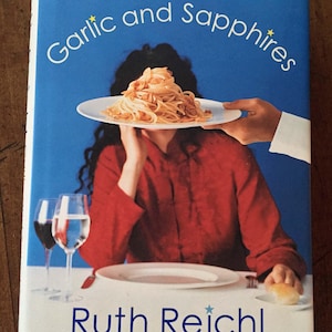 May include: Book cover for "Garlic and Sapphires" by Ruth Reichl. The cover features a woman in a red shirt with a plate of pasta held in front of her face. Two wine glasses and a plate are on the table. The background is blue.