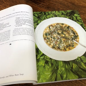 May include: A close-up of a white bowl filled with a white bean soup with escarole, parsnip, and red pepper flakes. The soup is garnished with grated cheese and is surrounded by a bed of fresh green lettuce.