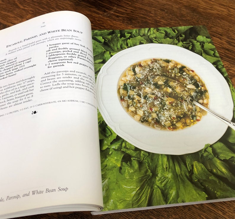 May include: A close-up of a white bowl filled with a white bean soup with escarole, parsnip, and red pepper flakes. The soup is garnished with grated cheese and is surrounded by a bed of fresh green lettuce.