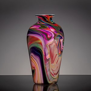 Puede incluir: Un jarrón de vidrio con un patrón arremolinado y jaspeado de colores vibrantes que incluyen morado intenso, rosa brillante, naranja y toques de verde. El recipiente tiene una superficie lisa y pulida con un cuerpo redondeado y una abertura estrecha.