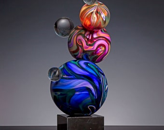May include: A glass sculpture with a black marble base. The sculpture is made of five glass orbs, each with a different swirling pattern of colors. The orbs are stacked on top of each other, with the largest orb at the bottom and the smallest orb at the top.