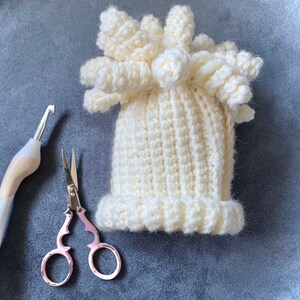 May include: A white crocheted hat with a fluffy pom-pom on top. The hat is lying on a gray surface with a pair of pink scissors and a crochet hook.