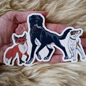 Può includere: Tre adesivi di animali: un cane nero, una volpe rossa e un cane bianco e marrone. Il cane nero e il cane bianco e marrone hanno collari turchese. Gli adesivi sono su una superficie strutturata marrone chiaro.