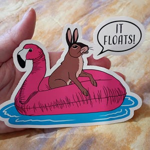 May include: A sticker featuring a brown rabbit riding a pink flamingo pool float in blue water. A speech bubble says "IT FLOATS!". The sticker is white with a die-cut edge.