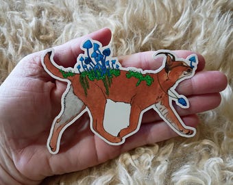 Op de afbeelding: Een gestanste sticker met een lichtbruine caracal met witte onderkant, versierd met vlekken groen mos en trossen felblauwe paddenstoelen op de rug. De caracal draagt een blauwe kristallen hanger om de nek.