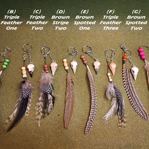 Op de afbeelding: Een verzameling handgemaakte sleutelhangers met veren, kleine schedelbedels en houten kralen. Elke sleutelhanger heeft een uniek veermotief, met variaties in kleur en patroon, waaronder bruine strepen en stippen.