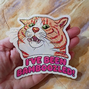 May include: A sticker with a cartoon cat design. The cat has orange and white fur, green eyes, and an open mouth. Below the cat is the text "I'VE BEEN BAMBOOZLED!" in pink. The sticker has a glittery, holographic finish.