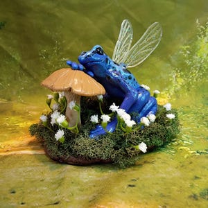 May include: A whimsical figurine of a blue frog with translucent wings, resting on a mushroom. The frog is speckled with black spots and sits amidst a bed of green moss and small white flowers. The mushroom has a tan cap.