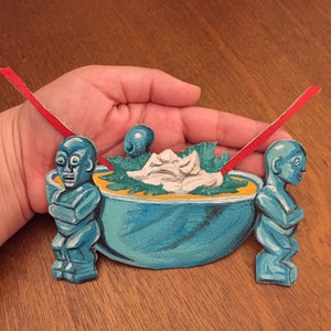 May include: A blue ceramic bowl with a yellow rim, filled with a white creamy substance and green leafy vegetables. Two blue tiki figures with red straws are on either side of the bowl.