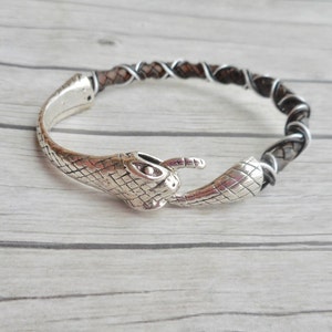 May include: A silver snake head bracelet with a brown leather strap. The bracelet is made of metal and has a snake head design. The leather strap is braided and has a black and white pattern.