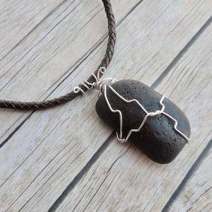 Puede incluir: Un colgante de piedra negra envuelto en alambre plateado, colgando de un collar de cordón de cuero marrón.