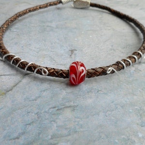 Puede incluir: Un collar de cordón de cuero marrón con un acento de alambre plateado y una cuenta de vidrio roja con remolinos blancos.