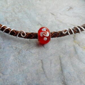 Puede incluir: Un brazalete de cuero marrón con detalles de alambre plateado y una cuenta de vidrio roja con un diseño de espiral blanco.