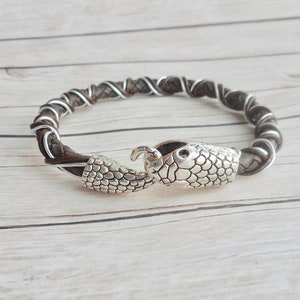 Op de afbeelding: Een bruin leren armband met een zilveren slangen sluiting. De armband is gevlochten met een zilveren draad.