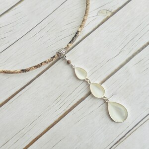 Peut inclure: Un collier avec un ras du cou à motif peau de serpent et un pendentif argenté. Le pendentif comporte trois pierres en forme de poire, blanc cassé. Le collier est présenté sur une surface en bois clair.