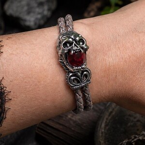 Può includere: Un bracciale in pelle intrecciata con un elemento centrale in metallo a forma di teschio, caratterizzato da una pietra preziosa rossa incastonata nella bocca. Il teschio presenta dettagli del viso e decorazioni metalliche su un cordino doppio.
