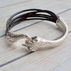 May include: A silver snake-shaped bracelet with a brown leather strap. The bracelet is made of metal and has a textured surface.
