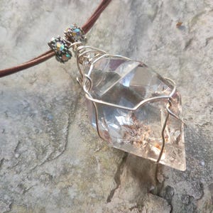 May include: A clear, faceted crystal pendant wrapped in silver-toned wire, suspended from a brown cord. The pendant features two small, iridescent, cylindrical beads at the top where the cord meets the wire-wrapped crystal structure.