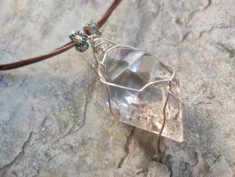 May include: A clear, faceted crystal pendant wrapped in silver-toned wire, suspended from a brown cord. The pendant features two small, iridescent, cylindrical beads at the top where the cord meets the wire-wrapped crystal structure.