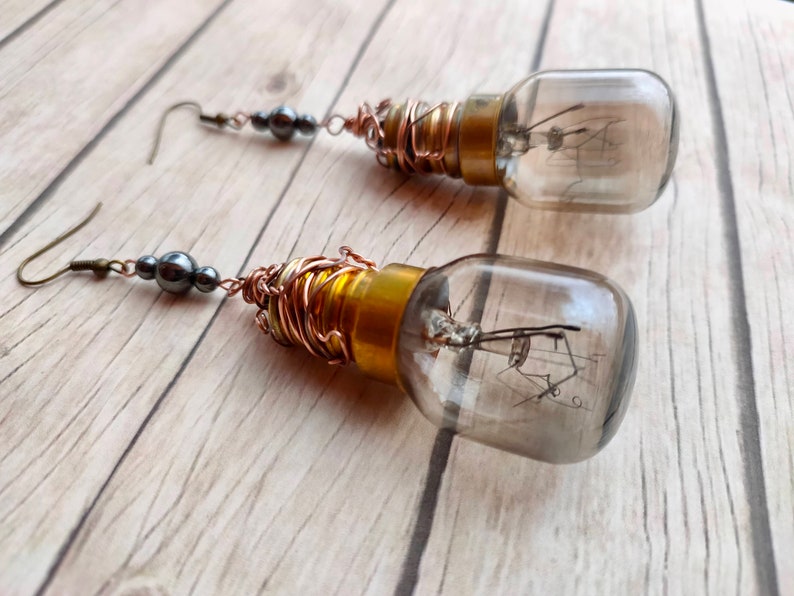 May include: A pair of earrings featuring small light bulbs wrapped in copper wire and accented with black beads. The bulbs are clear glass and have a gold-colored base.