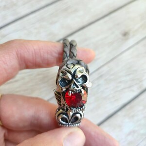 May include: A braided leather bracelet with a silver skull charm featuring a red gemstone. The skull has detailed features and a menacing expression.
