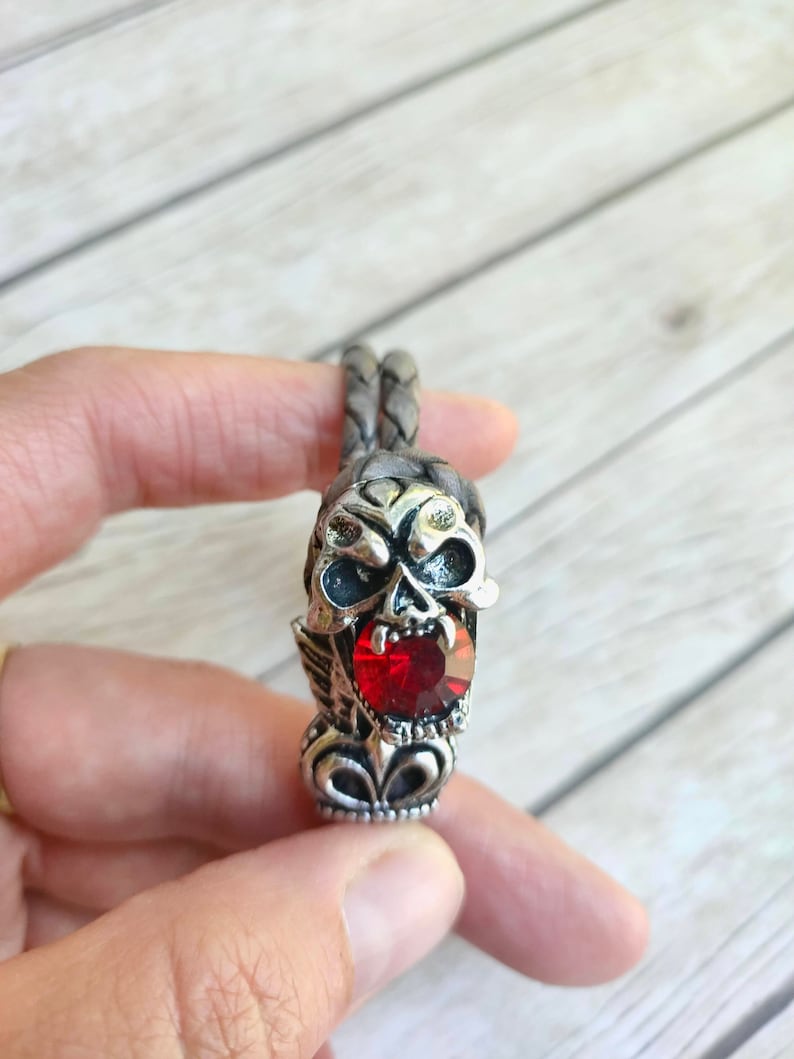 May include: A braided leather bracelet with a silver skull charm featuring a red gemstone. The skull has detailed features and a menacing expression.