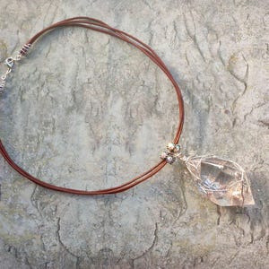 May include: A necklace featuring a clear, faceted crystal pendant encased in silver-toned wire wrapping. The pendant is attached to a brown cord that consists of three thin strands, secured with a silver-toned metal clasp and wire-wrapped accents.