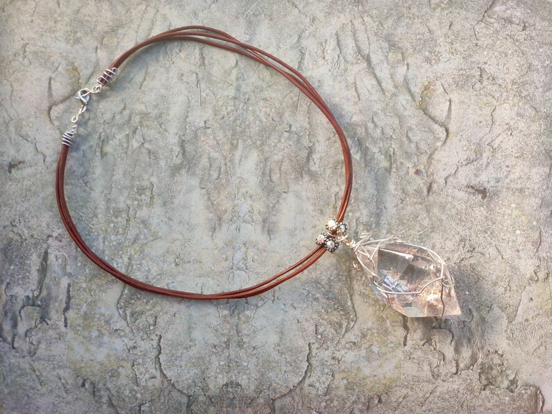 May include: A necklace featuring a clear, faceted crystal pendant encased in silver-toned wire wrapping. The pendant is attached to a brown cord that consists of three thin strands, secured with a silver-toned metal clasp and wire-wrapped accents.