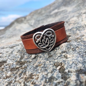 May include: A brown leather cuff bracelet with a silver Celtic knot heart charm. The bracelet is on a grey rock background.