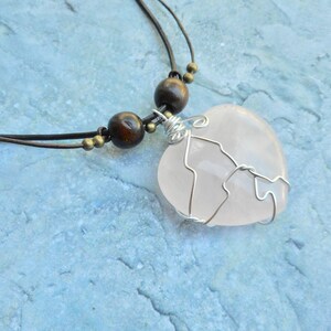 May include: A pink rose quartz heart pendant wrapped in silver wire, hanging from a brown leather cord with wooden beads.