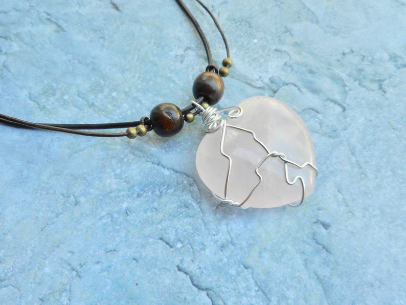 May include: A pink rose quartz heart pendant wrapped in silver wire, hanging from a brown leather cord with wooden beads.