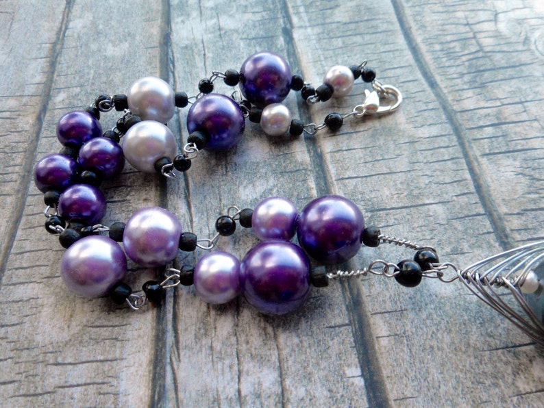 Peut inclure: Un collier en cha&icirc;ne argent&eacute;e avec des perles de couleur violette et blanche et des perles noires. Le collier a un fermoir argent&eacute; et un pendentif argent&eacute; avec un motif g&eacute;om&eacute;trique.