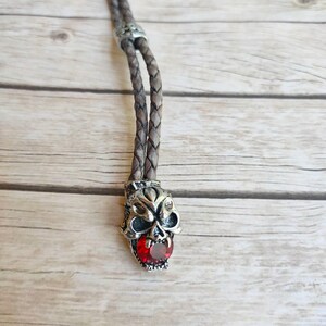May include: A brown leather bolo tie with a silver skull pendant featuring a red gemstone in its mouth.