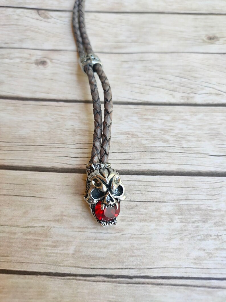 May include: A brown leather bolo tie with a silver skull pendant featuring a red gemstone in its mouth.