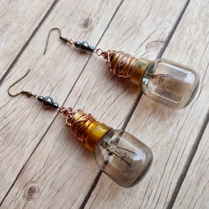 May include: A pair of long, dangling earrings featuring small, dark gray beads and a clear glass light bulb with a gold base wrapped in copper wire.
