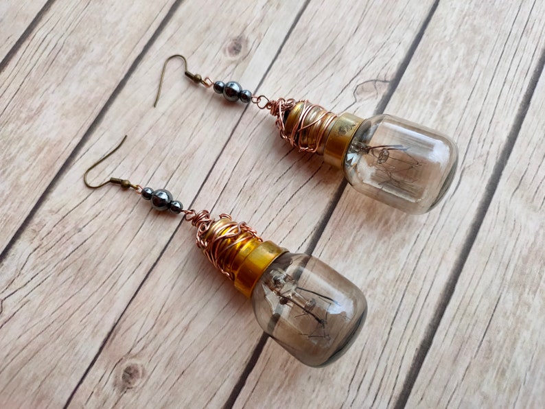 May include: A pair of long, dangling earrings featuring small, dark gray beads and a clear glass light bulb with a gold base wrapped in copper wire.