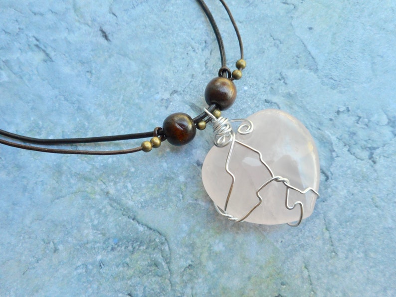 May include: A pink rose quartz heart pendant necklace with a silver wire wrap, brown wooden beads, and a brown leather cord.