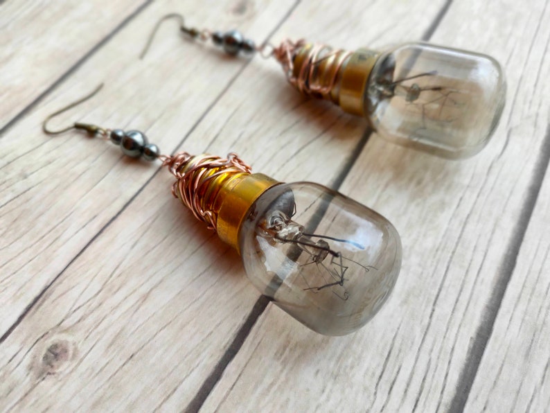 May include: A pair of earrings made from repurposed light bulbs. The bulbs are clear glass and have a copper wire wrapped around the base. The earrings are hanging from silver hooks with black beads.