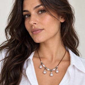 May include: A necklace featuring a dark brown cord adorned with five white spherical beads and small silver-toned metal spacers. The beads are arranged in a cascading pattern, with the center bead hanging slightly lower than the others.