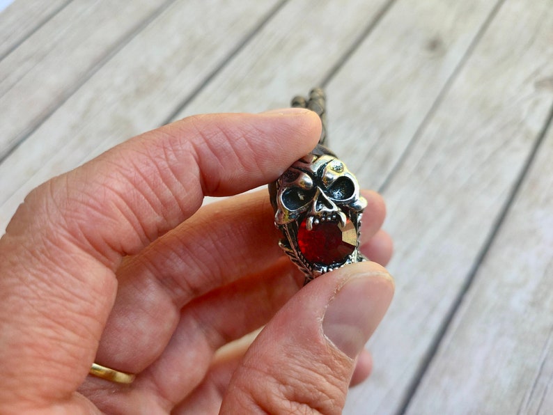 May include: A silver skull pendant with a red gemstone in the mouth. The pendant is on a leather cord.