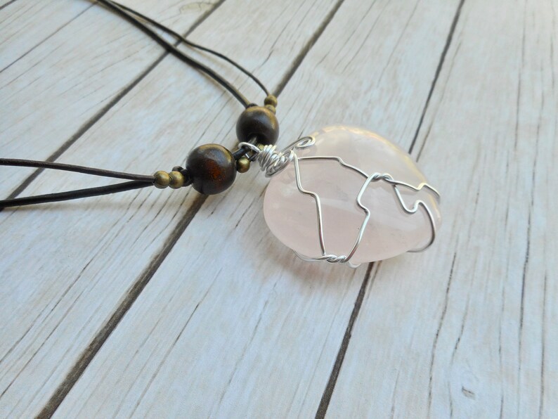 May include: A pink rose quartz heart pendant wrapped in silver wire, hanging from a brown leather cord with wooden beads.