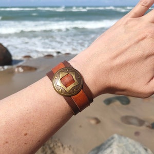 May include: A brown and tan leather cuff bracelet with a decorative brass centre. The bracelet features a geometric design in the centre and is worn on a wrist. The background shows a beach with waves.