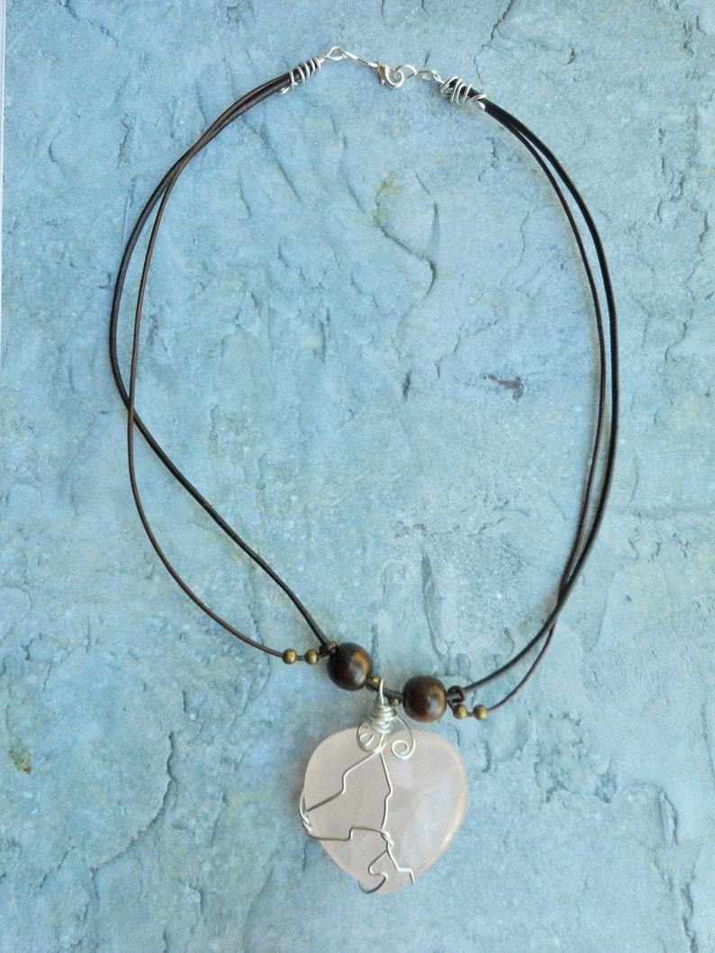 May include: A pink heart-shaped pendant necklace with a wire wrapped design. The necklace is made with brown leather cord and wooden beads.