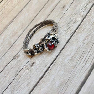 May include: A brown leather bracelet with a silver skull charm and a red gemstone. The bracelet is on a wooden surface.