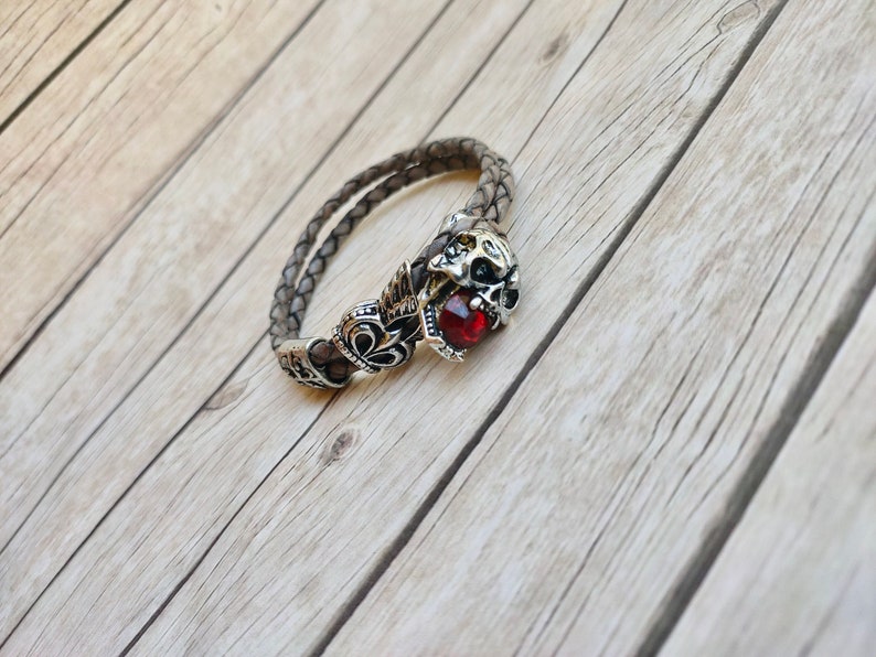 May include: A brown leather bracelet with a silver skull charm and a red gemstone. The bracelet is on a wooden surface.