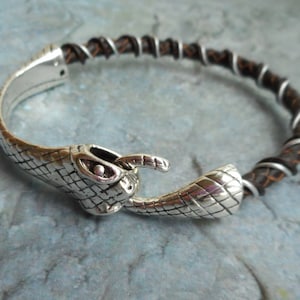May include: Silver bracelet featuring a snake head clasp. The band is brown leather, wrapped with silver-coloured wire. The snake head has a scale pattern and is detailed with eyes and a mouth.