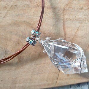 May include: A clear, faceted crystal pendant wrapped in silver-toned wire, suspended from a brown cord. The necklace features two small, cylindrical beads with iridescent, multi-coloured accents positioned above the crystal pendant.