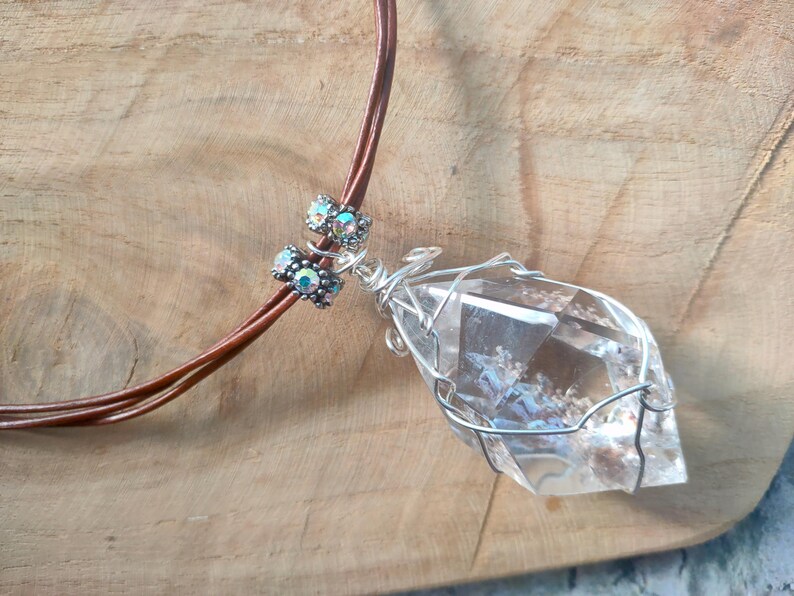May include: A clear, faceted crystal pendant wrapped in silver-toned wire, suspended from a brown cord. The necklace features two small, cylindrical beads with iridescent, multi-coloured accents positioned above the crystal pendant.